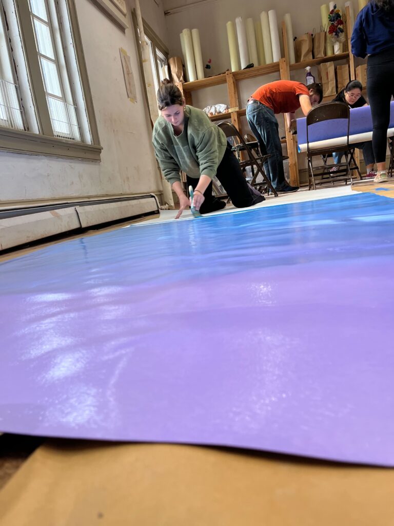 Emily Blaum kneels on the ground, painting the background of a large sign in a purple-to-blue gradient.