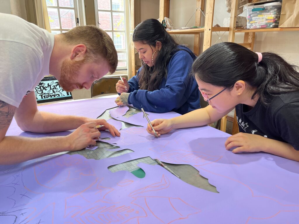 Eric Underwood, Yingying Lu, and Erika  Guadalupe Núñez, making cutouts in the shapes of leaves and birds in the purple sign.