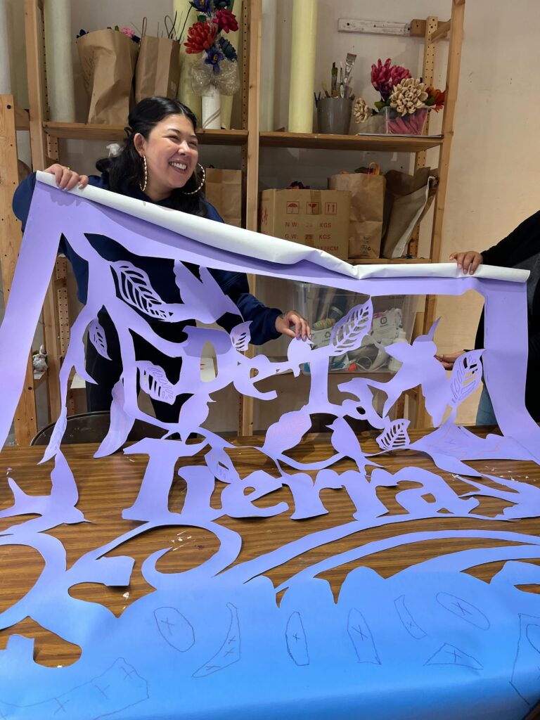 Erika Guadalupe Núñez smiles while holding up a purple and blue sign with cutouts in the shapes of birds and leaves and the words "De La Tierra Somos".