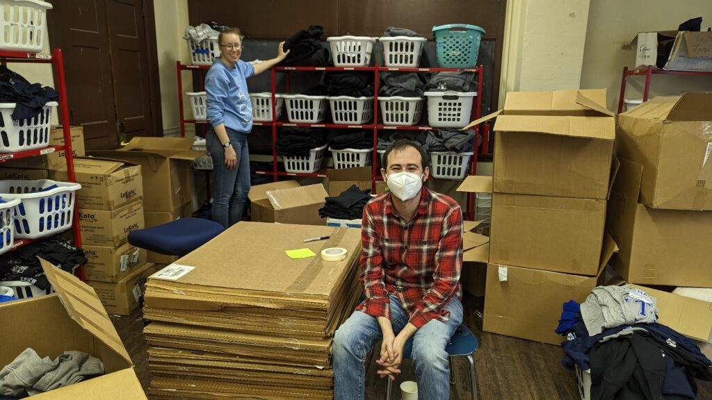 Melanie Bahti stands next to shelves of school uniforms, and Casey Whitman sits next to cardboard boxes.