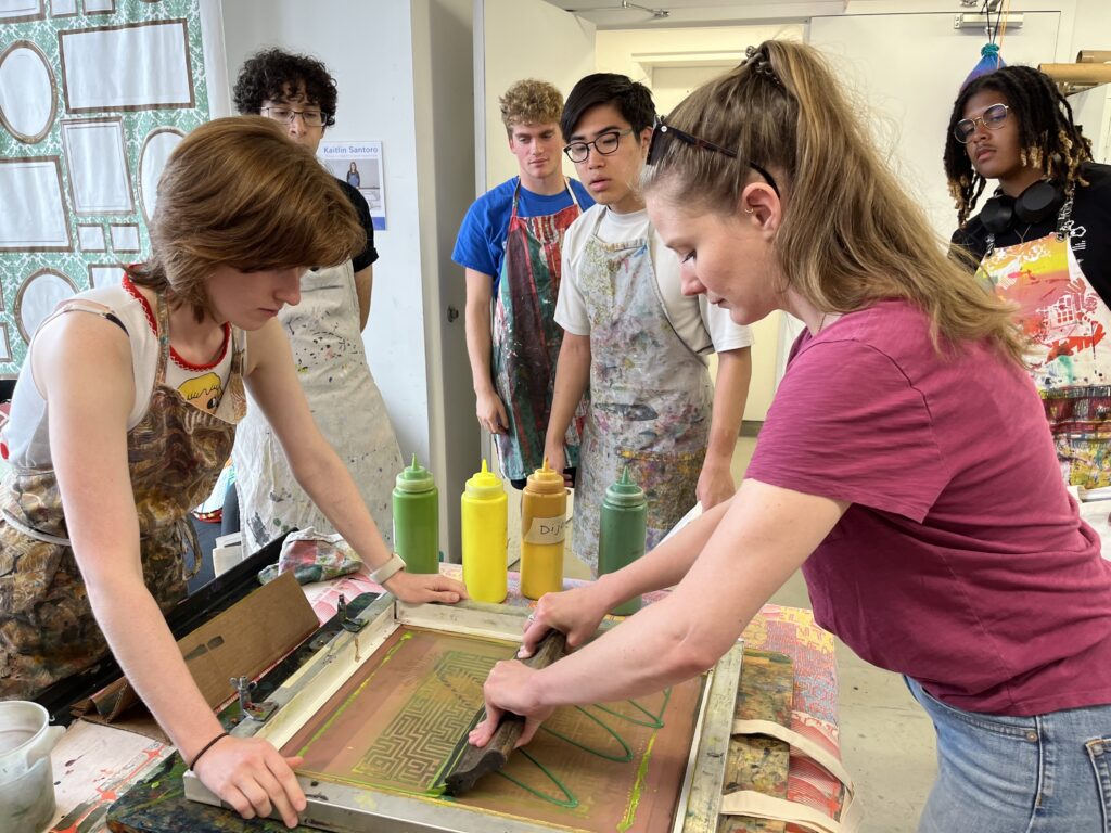 Stephanie Szymanski uses the squeegee to screen print a green and yellow design, while others watch in the background.