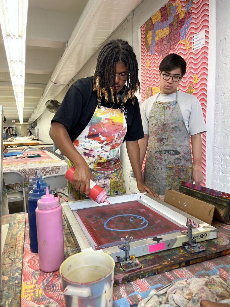 DJ Cintron adds red and blue fabric paint to the screen while Sebastian Castro watches.