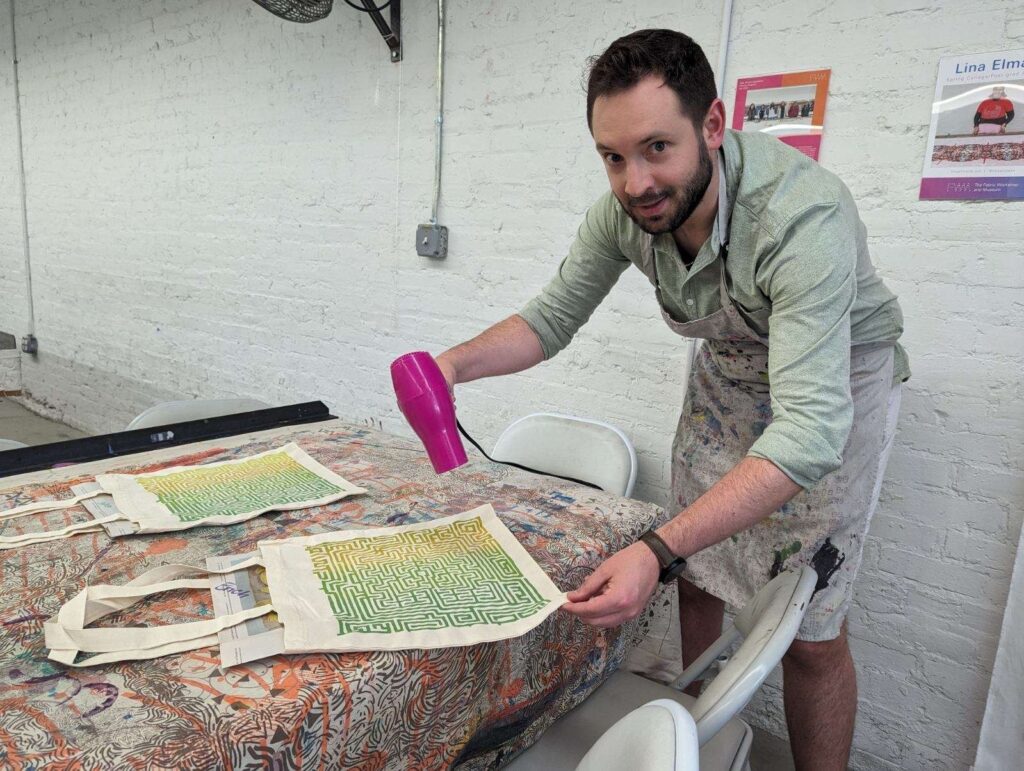 Erich Dress uses a hairdryer to dry the yellow and green design on his tote bag.