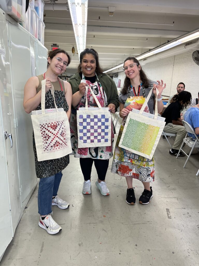 Emma Britez Ferrante, Nirali Patel, Shira Blady pose holding their tote bags. The designs of the bags resemble games: scrabble, checkers, and a maze.